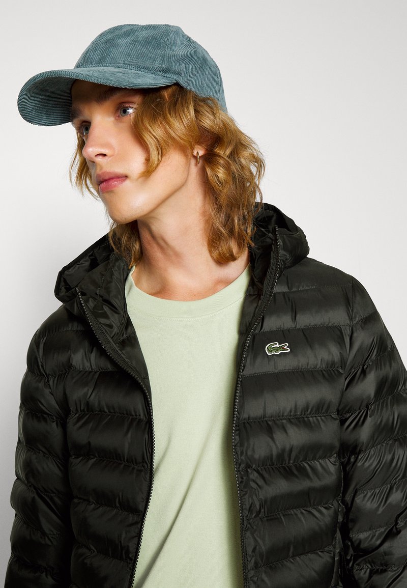 Doudoune noire avec un motif matelassé, t-shirt vert à col rond, et casquette en velours côtelé bleue, avec un petit logo brodé sur la doudoune.