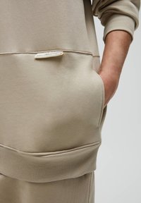 Sweatshirt beige à texture douce, avec une poche kangourou et des poignets côtelés. Une petite étiquette de marque est visible sur la poche.