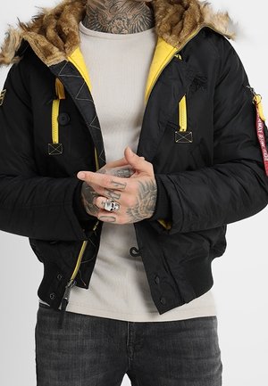 Veste bomber noire avec détails de fermeture éclair jaunes et capuche doublée de fourrure portée sur une chemise beige, montrant des mains tatouées avec des bagues.