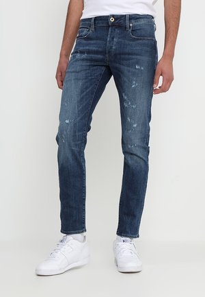 Jeans de mezclilla azul con corte ajustado, que presentan áreas desgastadas y detalles rasgados. Diseñados con cinco bolsillos y un cierre de botón.