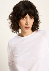 Frau mit welligem, dunkelbraunem Haar, die ein weißes Langarmshirt trägt und mit neutralem Ausdruck direkt in die Kamera schaut.