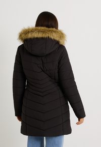 Femme portant un manteau matelassé noir mi-long avec capuche bordée de fourrure, debout de dos sur un fond uni.