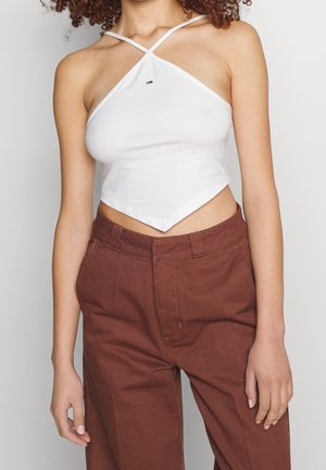 Femme portant un crop top blanc à col halter avec un petit logo et un pantalon marron taille haute, vue des épaules jusqu'aux hanches.
