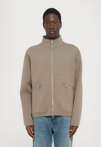 Chaqueta cremosa con cremallera y alto cuello, dos bolsillos frontales con cremallera y corte holgado. La tela parece suave con una textura blanda.