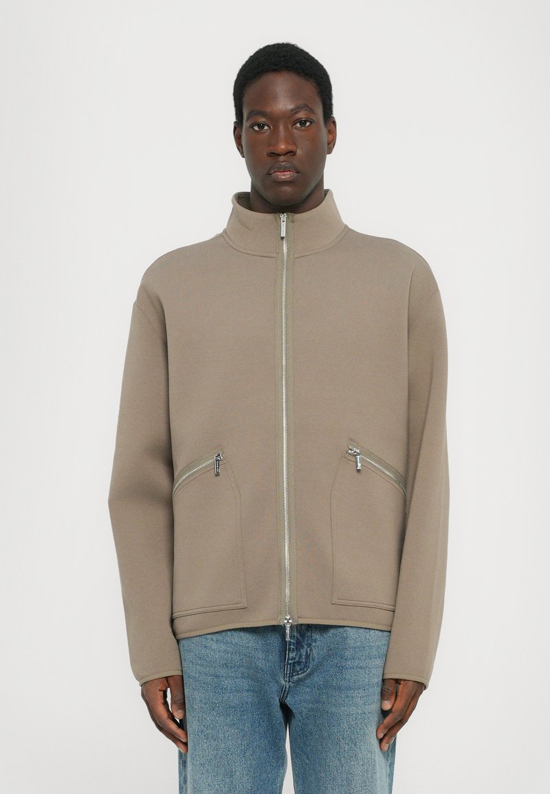 Chaqueta cremosa con cremallera y alto cuello, dos bolsillos frontales con cremallera y corte holgado. La tela parece suave con una textura blanda.
