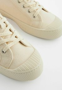 Baskets en toile beige avec bout rond, semelle en caoutchouc texturé et lacets plats. Présente des œillets en métal et une tige en tissu lisse.