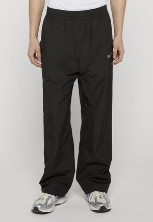 Mand iført løse sorte træningsbukser og hvide sneakers foran en ensfarvet grå baggrund, hænderne hvilende langs siderne.