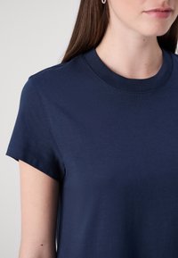 Vrouw in een marineblauwe t-shirt met korte mouwen en parel oorbelletjes, afgebeeld van de nek tot het bovenste deel van de torso tegen een lichte achtergrond.