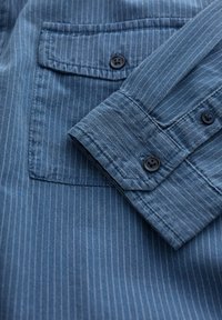 Blau gestreift, kurzärmliges Hemd mit einer Brusttasche, schwarzen Knöpfen und strukturiertem Stoff. Die Streifen sind vertikal ausgerichtet und dezent.