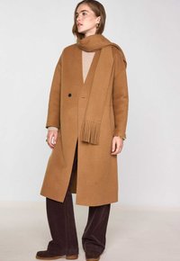 Lungo cappotto color cammello con texture liscia, dotato di chiusura con un solo bottone, tasche laterali e una sciarpa frangiata abbinata. Abbinato a pantaloni in corduroy scuri.