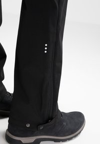 Pantalons étanches noirs avec zips latéraux, détail de trois points blancs, associés à des bottes en suede noires avec des accents gris et une semelle texturée.