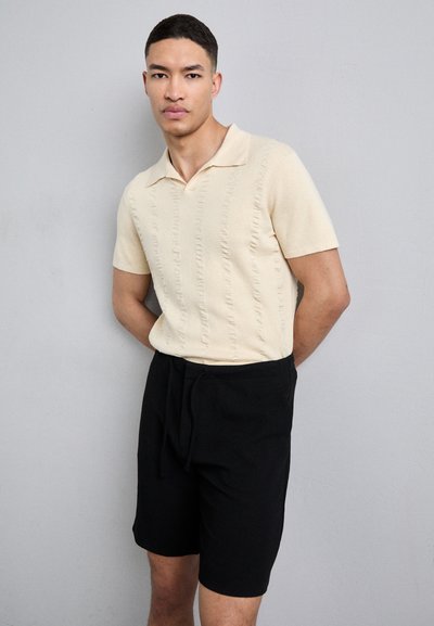 Beige kortærmet polo-shirt med teksturerede lodrette linjer, parret med sorte shorts med elastisk talje og snøre.