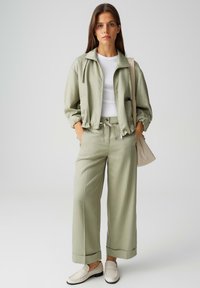 Femme debout, les mains dans les poches, portant une veste et un pantalon vert sauge, une chemise blanche, des mocassins crème, et tenant un sac fourre-tout beige.