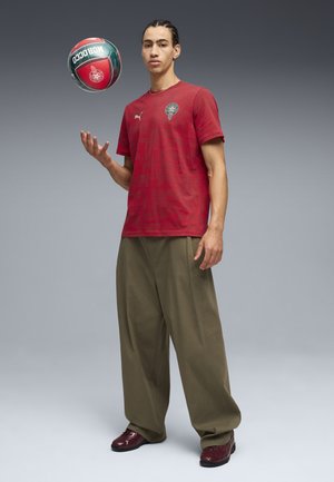 Jeune homme en chemise de sport rouge et pantalon olive faisant tournoyer un ballon de football rouge et vert sur une seule main de manière décontractée.