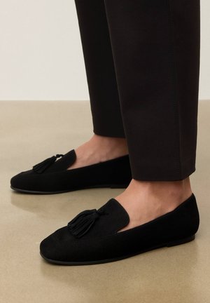 Mocasines de ante negro con borlas usados por una persona de pie sobre un suelo liso beige, combinados con pantalones negros ajustados.