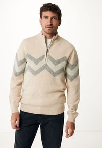 Beige zip-kraag trui met een chevronpatroon in lichtgrijs en crème. Gebreide textuur met ribbelmanchetten en -zoom. Gecombineerd met donkere spijkerbroeken.
