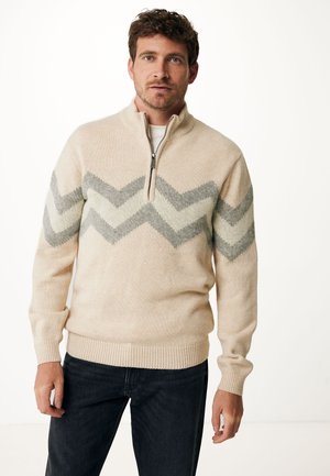 Beige zip-kraag trui met een chevronpatroon in lichtgrijs en crème. Gebreide textuur met ribbelmanchetten en -zoom. Gecombineerd met donkere spijkerbroeken.