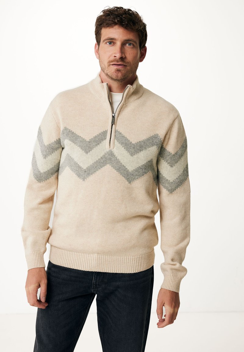 Beige zip-kraag trui met een chevronpatroon in lichtgrijs en crème. Gebreide textuur met ribbelmanchetten en -zoom. Gecombineerd met donkere spijkerbroeken.