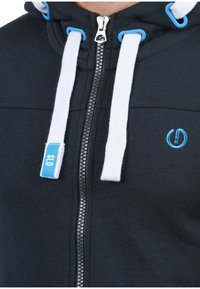 Marineblaues Zip-Hoodie aus weichem Stoff, mit weißen Kordeln, einem silbernen Reißverschluss und einer blauen Logo-Akzentuierung.