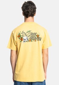 Gele katoen t-shirt met korte mouwen, met een doodskop- en bloempatroon op de achterzijde, en de tekst "DC Shoe Company".