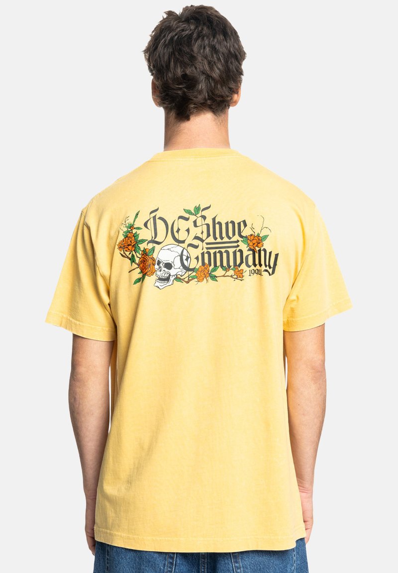 Gele katoen t-shirt met korte mouwen, met een doodskop- en bloempatroon op de achterzijde, en de tekst "DC Shoe Company".