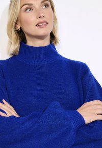 Moteris su šviesiais plaukais, vilkinti mėlyną megztinį su apykakle, sukryžiavusi rankas, žiūri į viršų ir šiek tiek į šoną prieš baltą foną.