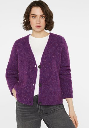 Frau trägt eine lila strukturierte Strickjacke mit Knöpfen über einem weißen Hemd und dunkelgrauen Jeans, steht vor einem schlichten weißen Hintergrund.