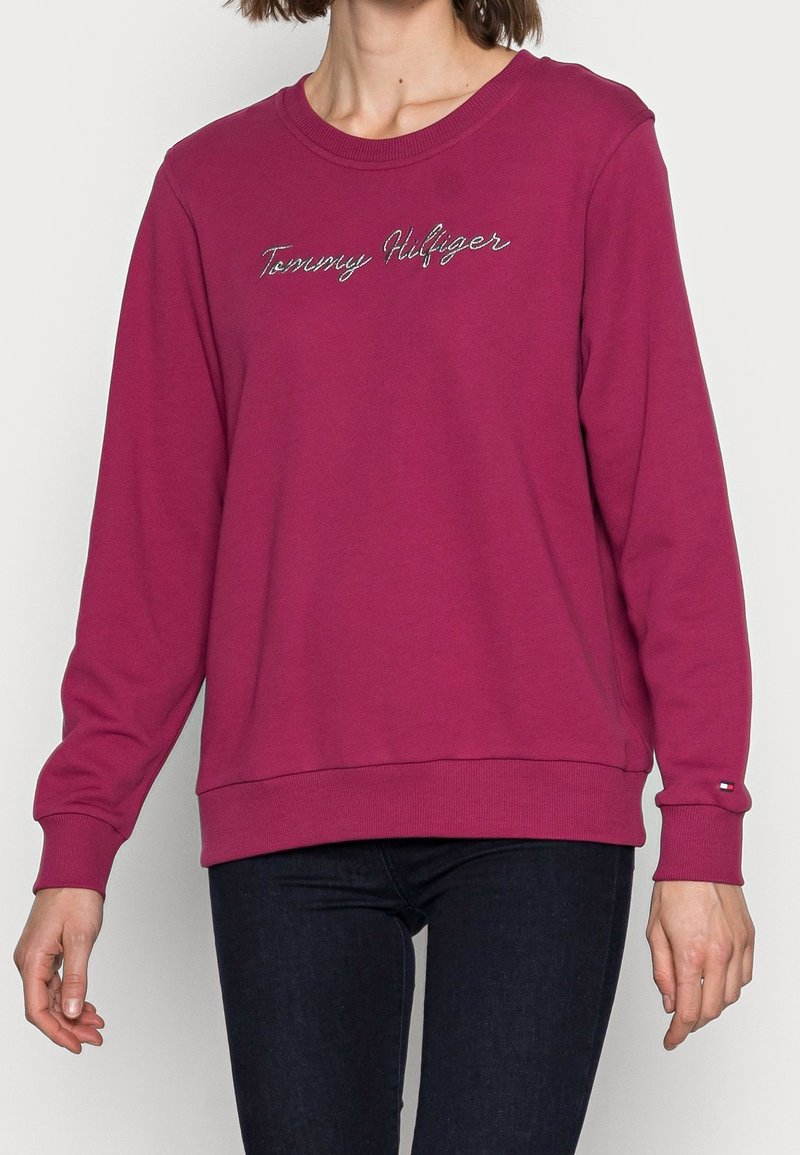 Femme portant un sweat-shirt magenta à manches longues Tommy Hilfiger et un jean foncé, debout devant un fond clair uni.