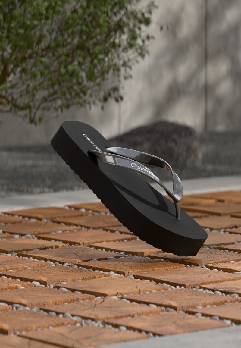 Μαύρο σαγιονάρι Calvin Klein με λαμπερές λουρίδες, τοποθετημένο σε ξύλινο πλακάκι σε εξωτερικό χώρο, κοντά σε θολό δέντρο και τοίχο.