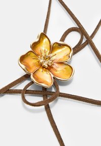 Broška v obliki zlate in oranžne rože z osrednjim kristalom iz rhinestona, nameščena na rjavih velurjih. Cvetlični motiv ima gladko, sijočo teksturo.