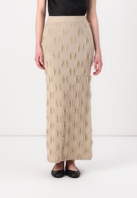 Beige, knielanges Strickrock mit strukturiertem Fransenmuster, tailliertem Bund und geripptem Saum. Getragen mit schwarzen flachen Schuhen und einem schwarzen Oberteil.