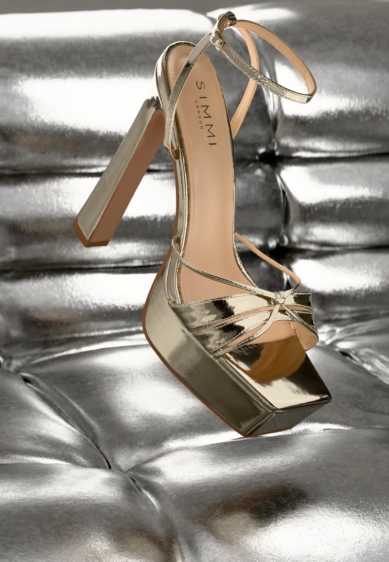 Scarpa con tacco alto in metallo dorato, con punta quadrata, apertura frontale e sottile cinturino alla caviglia, appoggiata su uno sfondo silver imbottito.