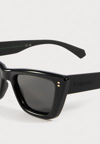 Svart plastsolbriller med firkantede linser, med en jevn overflate, to gullfargede prikkene på fronten, og en matte svart arm med merke.