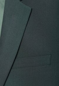 Dunkelgrüner Anzugstoff mit Fischgrätenmuster, glatter Oberfläche und einer schlichten, eleganten Brusttasche. Maßgeschneiderte Gestaltung mit scharfen Revers.