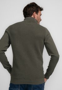 Olivgrüner Pullover mit strukturiertem Strickdesign, hohem Kragen und gerippten Bündchen und Saum. Tragen von einer Person, die seitlich steht.