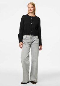 Cárdigan negro de punto con escote redondo y seis botones de perla. Presenta mangas largas con lazos en los puños. Combinado con jeans de pierna ancha en gris claro.