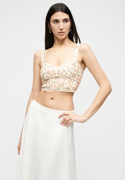 Γυναίκα με μακριά σκούρα μαλλιά φοράει ένα ανθισμένο κοντό τοπ με τιράντες και μια λευκή φούστα με ψηλή μέση, ποζάροντας με το ένα χέρι στη μέση.