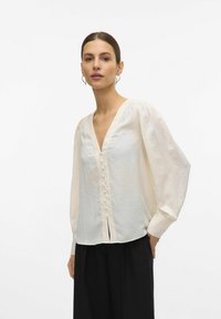 Blouse légère de couleur crème avec un décolleté en V, des manches bouffantes et un plastron décoratif à boutons. Associée à un pantalon noir. Design simple.