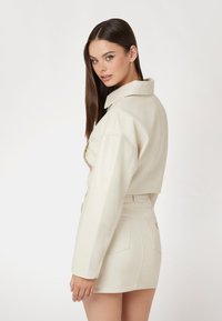 Nupjautas kreminės spalvos odinis švarkas su plačiu apykakle, šoninėmis kišenėmis ir suderinta aukštu liemeniu mini sijonu. Lygus paviršius, minimalus dekoro elementų kiekis.