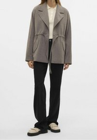 Grijze fleecejas met een taillekoord, oversized revers en twee voorkanten zakken, gecombineerd met zwarte broek en beige schoenen.