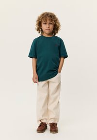 Σκούρο πετρόλ μπλουζάκι με κοντά μανίκια και διακριτικό λογότυπο στο στήθος, συνδυασμένο με εκρού παντελόνι φαρδιάς γραμμής και καφέ παπούτσια.