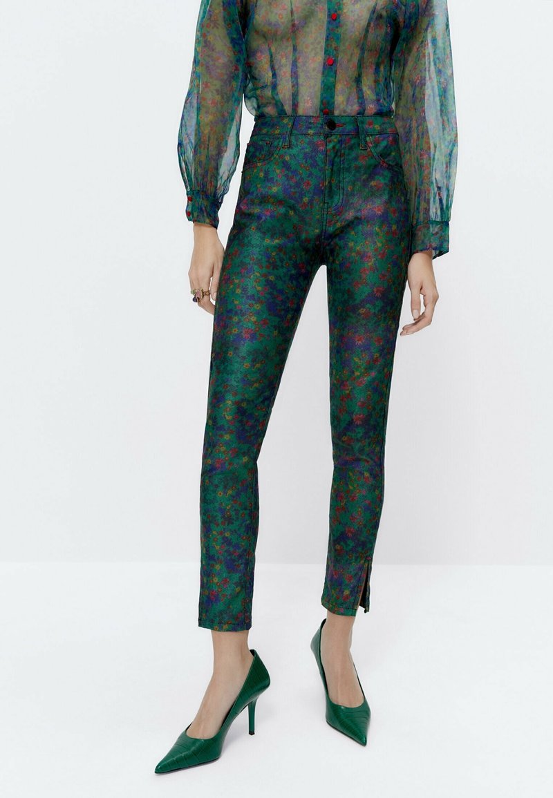 Zielone spodnie w kwiatowy wzór z błyszczącym wykończeniem i zamkiem błyskawicznym na dole, w parze z zielonymi szpilkami w wzory oraz przezroczystą bluzką z długim rękawem.