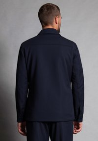 Navy blue, long-sleeve jacket with a flat collar, side seams, and buttoned cuffs, made from a smooth fabric with a structured design.