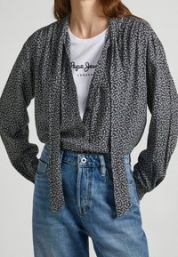 Blusa negra con estampado y lazo frontal, de mangas largas y corte relajado, combinada con una camiseta gráfica blanca y jeans de cintura alta azules.