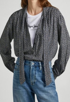 Blouse noire à motifs avec un nœud à l'avant, manches longues et coupe décontractée, superposée sur un t-shirt graphique blanc et un jean taille haute bleu.