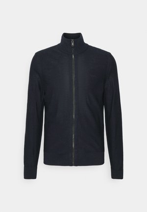 Suéter de punto azul marino con cierre, cuello alto, puños y cinturilla acanalados, con un sutil logo en el pecho. Textura suave, diseño ajustado.