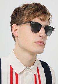 Ochelari de soare Ray-Ban negri cu lentile închise, cadru subțire și design unghiular elegant, purtați de o persoană în cămașă cu dungi deschise.
