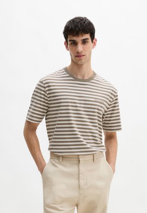 Junger Mann trägt ein beige-weiß gestreiftes kurzärmeliges Hemd und hellbeige Hose, steht mit den Händen in den Taschen vor weißem Hintergrund.