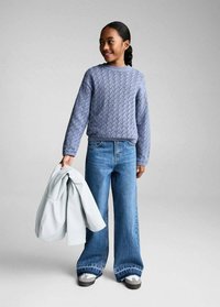 Helles blaues Strickpullover mit strukturiertem Muster, kombiniert mit weit geschnittenen blauen Jeans. Das Modell hält eine hellgraue Jacke und trägt graue Sneakers.