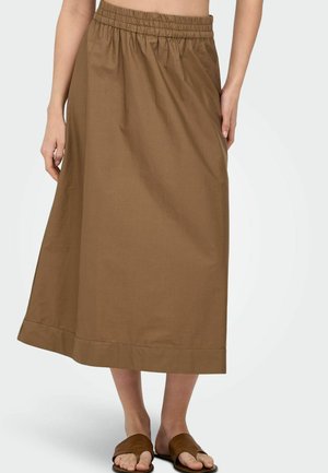 Femme portant une jupe marron longue jusqu'aux chevilles avec une ceinture élastique et des sandales plates ouvertes marron, debout devant un fond blanc.